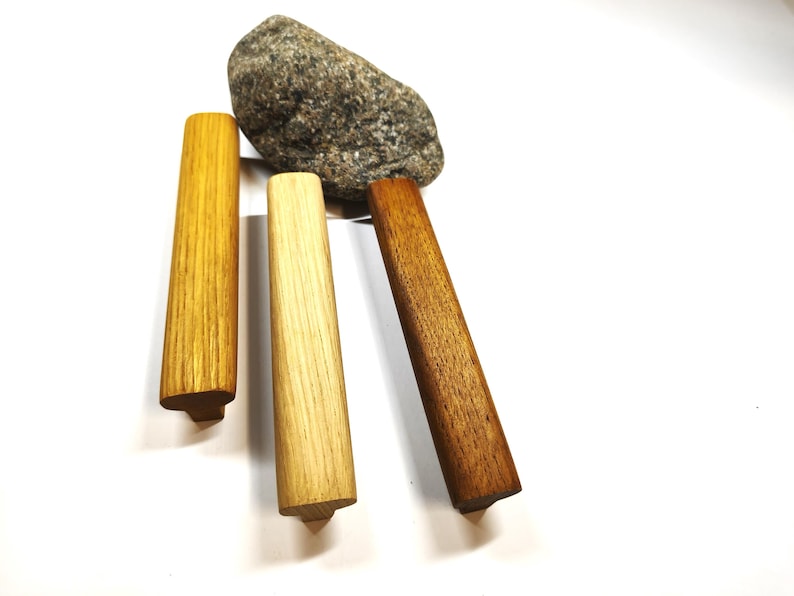 May include: Three elongated wooden cabinet pulls in varying shades of light, medium, and dark brown wood grain, positioned vertically near a single grey speckled stone. Each handle features a T-shaped profile with a flat top and a narrow base.