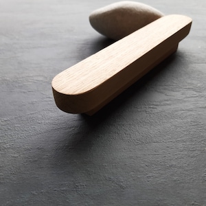 May include: A wooden drawer pull with a rounded rectangular shape. The pull is made of light brown wood and has a smooth finish.