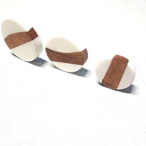May include: Three oval-shaped earrings with a white background and a brown wood inlay. The wood inlay is a different shape and size on each earring.