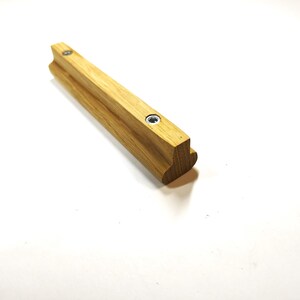May include: A light brown wooden cabinet handle with a T-shaped profile and two circular metal screw inserts embedded along the top surface. The handle features a smooth, natural wood grain finish and a rounded edge on the bottom side.