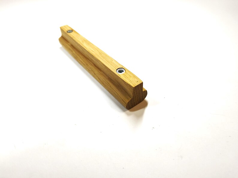 May include: A light brown wooden cabinet handle with a T-shaped profile and two circular metal screw inserts embedded along the top surface. The handle features a smooth, natural wood grain finish and a rounded edge on the bottom side.