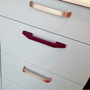 Könnte beinhalten: Weiße Schrank Schubladen mit drei verschiedenen Griffen. Die oberste Schublade hat einen silbernen Griff, die mittlere Schublade hat einen burgunderfarbenen Griff und die unterste Schublade hat einen goldenen Griff.