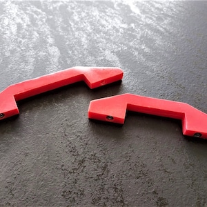 May include: Two red plastic handles with a rounded rectangular shape. Each handle has a small hole on either side.