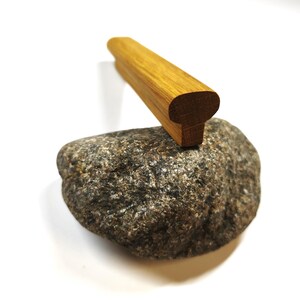 May include: A smooth, elongated piece of light brown wood with a rounded top and a flat base, resting on top of a textured, grey and black speckled natural stone. The wooden object features a visible grain pattern and a uniform, rectangular shape.