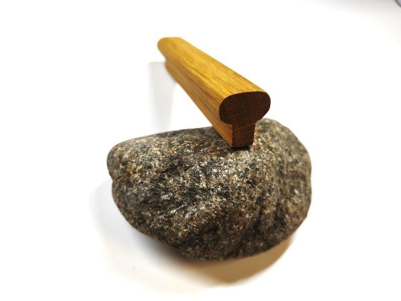 May include: A smooth, elongated piece of light brown wood with a rounded top and a flat base, resting on top of a textured, grey and black speckled natural stone. The wooden object features a visible grain pattern and a uniform, rectangular shape.