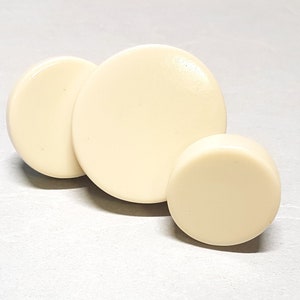 Puede incluir: Tres botones redondos de diferentes tamaños, de color blanco roto.