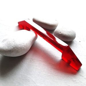 Peut inclure: Une poignée en verre de couleur rouge vif en forme de flèche avec une finition lisse et polie. La poignée repose sur trois pierres blanches lisses.