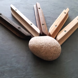 Op de afbeelding: Zes houten wandhaken in verschillende houttinten, waaronder naturel, bruin en wit, gerangschikt in een waaier rond een lichtbruine steen. Elke haak heeft twee zichtbare schroeven.