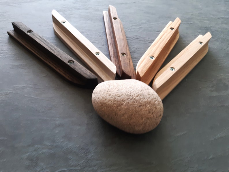Op de afbeelding: Zes houten wandhaken in verschillende houttinten, waaronder naturel, bruin en wit, gerangschikt in een waaier rond een lichtbruine steen. Elke haak heeft twee zichtbare schroeven.