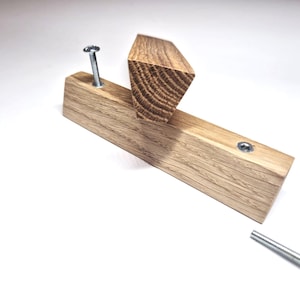 May include: A light brown wooden wall hook with a triangular profile and visible wood grain. The hook features a horizontal base with a vertical projection, accompanied by two silver metal screws for installation, one inserted and one separate.