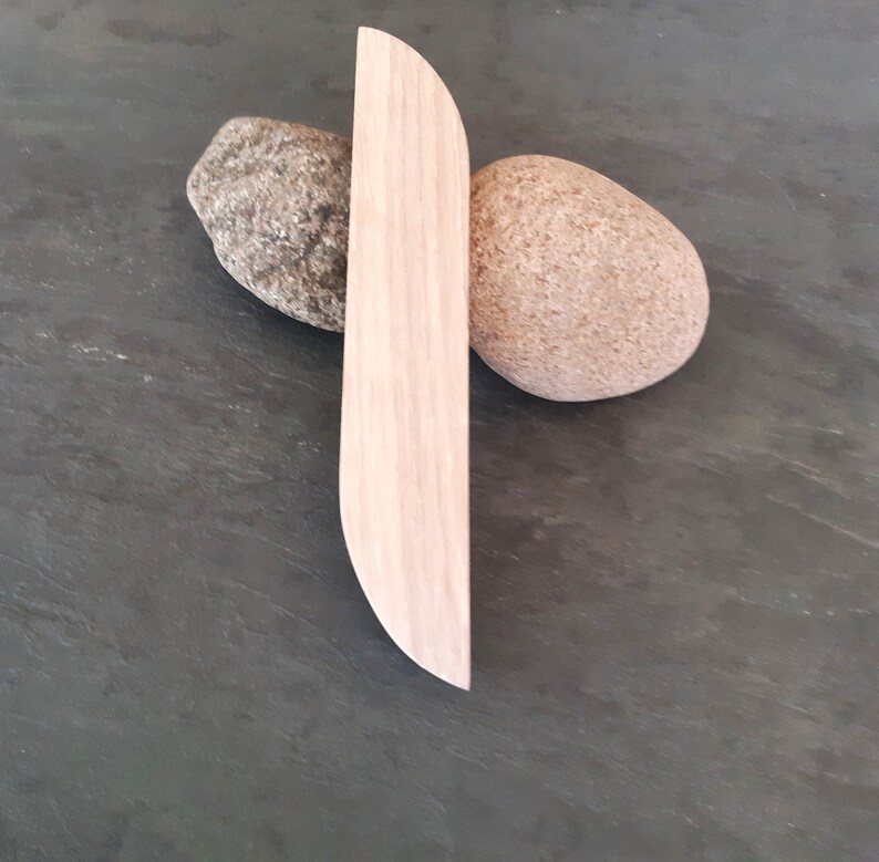 Op de afbeelding: Een lichtbruine houten carving in de vorm van een lang, dun, gebogen blad. De carving rust op twee ronde, grijze en bruine stenen.