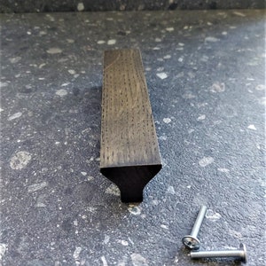 Puede incluir: Un tirador de armario de madera marrón oscuro con una forma rectangular ligeramente cónica y un borde biselado. El tirador tiene un acabado oscuro y un sutil patrón de veta de madera. Se incluyen dos tornillos plateados.