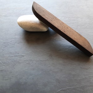 Peut inclure: Un objet en bois brun foncé avec un bord arrondi, reposant sur une pierre blanche. L'objet est sur une surface texturée grise.