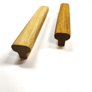 Op de afbeelding: Twee lichtbruine houten meubelgrepen met een glad, langwerpig T-vormig profiel. De handgrepen hebben een zichtbaar natuurlijk houtnerfpatroon en een afgerond bovenoppervlak, ontworpen voor bevestiging aan meubeldeuren of lades.