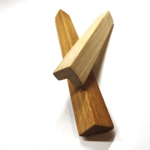 Puede incluir: Dos protectores de esquina de madera en forma de L con acabado liso. Una pieza presenta una veta de madera natural de color beige claro, mientras que la otra muestra un tono marrón cálido más profundo. Ambas tienen un extremo puntiagudo.