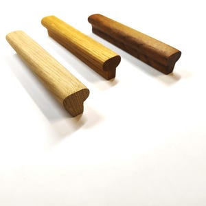 May include: Three linear wooden cabinet pulls arranged diagonally. Each handle features a smooth, elongated rectangular top with a rounded profile and a solid base. The set displays three distinct wood tones: light oak, medium oak, and dark walnut.