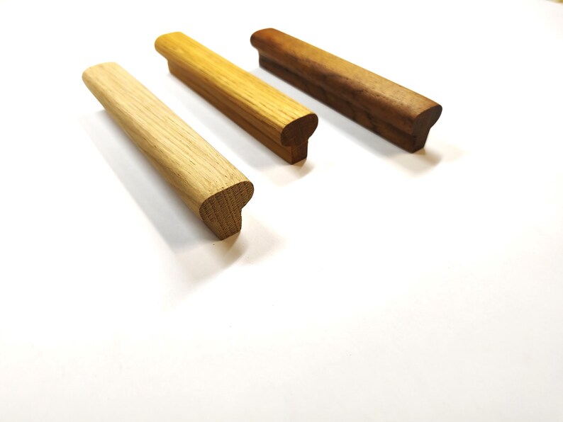 May include: Three linear wooden cabinet pulls arranged diagonally. Each handle features a smooth, elongated rectangular top with a rounded profile and a solid base. The set displays three distinct wood tones: light oak, medium oak, and dark walnut.