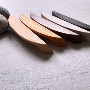 May include: Six wooden cabinet pulls in various shades of wood, from light to dark, arranged in a row on a white textured surface.