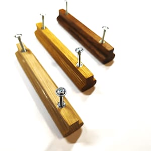 May include: Three linear wooden drawer pulls with a smooth finish, each featuring two silver metal screws inserted into the top. The pulls vary in wood grain color, ranging from a light golden tan to a deeper, richer brown tone, arranged diagonally.