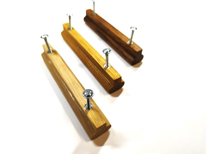 May include: Three linear wooden drawer pulls with a smooth finish, each featuring two silver metal screws inserted into the top. The pulls vary in wood grain color, ranging from a light golden tan to a deeper, richer brown tone, arranged diagonally.
