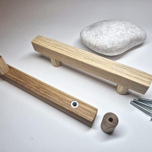 Può includere: Due maniglie per armadietti in legno chiaro con profilo rettangolare, una assemblata con una vite in metallo e una smontata con un distanziatore cilindrico separato e tre viti in metallo aggiuntive, accanto a una pietra grigia liscia.