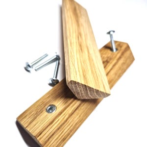 Könnte beinhalten: Zwei rechteckige, hellbraune Holzgriffe für Schränke mit sichtbarer Holzmaserung. Ein Griff liegt flach mit einem silbernen Metalleinsatz für Schrauben, während der andere aufrecht steht. Drei separate silberne Metallschrauben liegen bei.