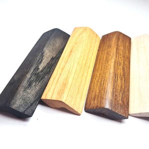 May include: Four wooden drawer pulls with a triangular profile, displayed in a row. The finishes include a dark charcoal stain, a light natural wood, a medium brown wood tone, and a pale unfinished wood. Several silver metal screws are included.