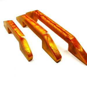 Può includere: Tre maniglie per armadietto di diverse lunghezze, ognuna caratterizzata da un motivo marmorizzato traslucido arancione e giallo. Le maniglie presentano una finitura liscia e lucida con un design angolare distinto, mostrando una vibrante miscela di colori.