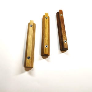May include: Three rectangular wooden cabinet pulls arranged vertically. Each pull features a distinct wood grain pattern, ranging from light to dark brown, and includes two circular metal screw holes for mounting purposes on furniture.