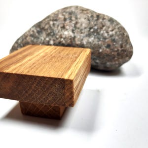 May include: A wooden knob with a square top and a rounded base. The knob is made of light brown wood with visible grain. The knob is sitting on a white surface.