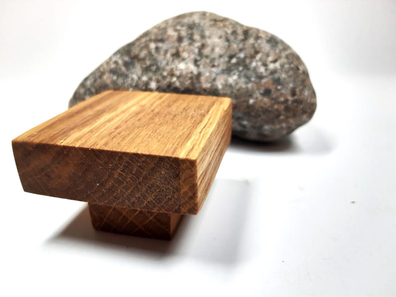 May include: A wooden knob with a square top and a rounded base. The knob is made of light brown wood with visible grain. The knob is sitting on a white surface.