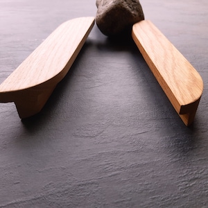 Pode incluir: Duas puxadores de armário de madeira com forma retangular arredondada. Os puxadores são de madeira castanho claro com um padrão de grão natural.