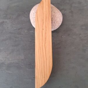 Op de afbeelding: Een houten, lichtbruin, rond, mesvormig object met een glad oppervlak en een licht gebogen rand. Het object ligt op een grijs, getextureerd oppervlak.
