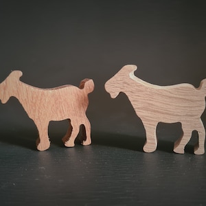 Pode incluir: Duas silhuetas de cabra de madeira, uma virada para a esquerda e a outra para a direita, estão em pé sobre uma superfície escura. As cabras são feitas de madeira marrom claro e têm um design simples e estilizado.
