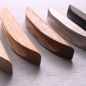 May include: Five wooden cabinet pulls in different finishes, including natural wood, brown, white, and black. The pulls are all curved and have a simple, modern design.