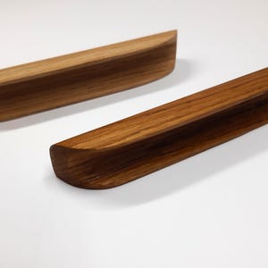 May include: Two wooden drawer pulls with a natural wood finish. The pulls are curved and have a slight lip on the top and bottom.