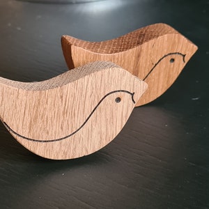 Peut inclure: Deux ornements d'oiseaux en bois avec un design simple et sculpté. Les oiseaux sont en bois brun clair et ont une finition naturelle.
