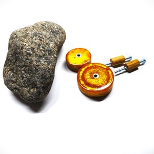 May include: A natural grey speckled stone positioned next to two circular amber-colored resin drawer knobs. Each knob features a central metal screw hole and is accompanied by a small wooden cylindrical spacer and a silver metal mounting bolt.