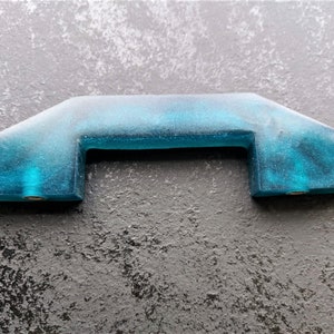 Puede incluir: Una manija de puerta de armario de resina azul turquesa con un diseño único ligeramente curvado.