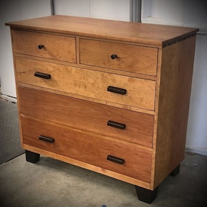 May include: A wooden dresser with five drawers and a top surface. The dresser is made of light and dark brown wood with black drawer pulls and feet. The top two drawers are smaller than the bottom three.