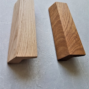 Puede incluir: Dos tiradores de puerta de armario de madera, uno de madera clara y otro de madera oscura. Ambos tienen una forma rectangular con una ligera curva en la parte inferior.