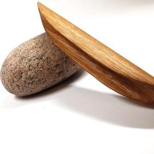 Può includere: Un oggetto di legno a forma di barca con fondo arrotondato e superficie liscia e lucida. L'oggetto è in legno marrone chiaro e presenta una venatura naturale. È appoggiato su una roccia grigia testurizzata.