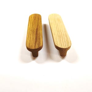 Op de afbeelding: Twee langwerpige, ovaalvormige houten meubelgrepen of ladehandgrepen. De ene greep heeft een mediumbruine houtnerfafwerking, terwijl de andere een lichtere, bleke houtnerf vertoont. Beide items hebben een glad, afgerond bovenoppervlak.
