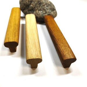May include: Three T-shaped wooden handles with distinct wood grain patterns and varying brown tones, ranging from a light golden yellow to a medium tan and a deeper reddish brown, arranged vertically with a grey stone positioned behind them.