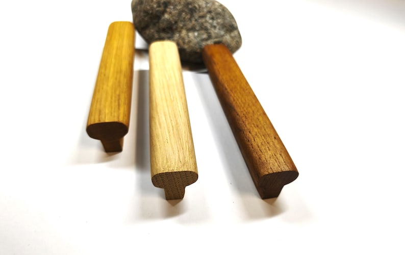 May include: Three T-shaped wooden handles with distinct wood grain patterns and varying brown tones, ranging from a light golden yellow to a medium tan and a deeper reddish brown, arranged vertically with a grey stone positioned behind them.