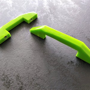 Puede incluir: Dos tiradores de armario verde brillante con un diseño geométrico. Cada tirador tiene un agarre rectangular y soportes angulados, con orificios para tornillos visibles. Los tiradores son de un material sólido y liso, sobre un fondo gris oscuro.