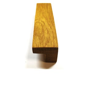 May include: A rectangular cabinet pull handle crafted from light brown oak wood with a visible natural grain pattern. The handle features an L-shaped profile with a flat top surface and a downward-facing edge, designed for mounting onto furniture.