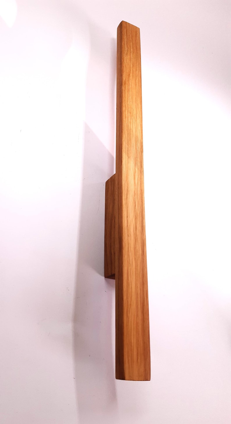 May include: A wooden wall sconce with a natural wood finish. The sconce has a rectangular shape and is mounted to the wall with a small, rectangular bracket.
