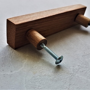 Op de afbeelding: Een houten ladegreep met een rechthoekige vorm en twee ronde knoppen. De greep is gemaakt van lichtbruin hout en heeft een natuurlijke afwerking. De knoppen zijn met schroeven aan de greep bevestigd.