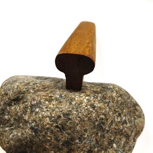 May include: A smooth, elongated wooden handle with a T-shaped profile, featuring a warm brown grain pattern. The handle has a rounded top section that tapers down into a narrower base, showcasing a polished finish and natural wood texture throughout.